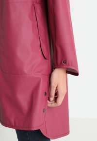 Wodoodporny bordowy płaszcz o gładkiej fakturze, z zatrzaskami, boczną kieszenią i długimi rękawami, prezentujący dopasowaną sylwetkę.