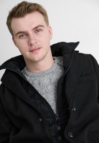 Schwarze Wollmischungjacke mit einem Reißverschluss und großem Kragen, über einem hellgrauen Strickpullover mit Zopfmuster getragen.