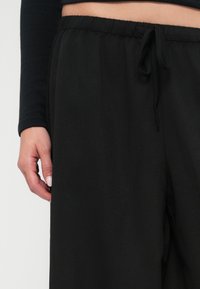 Mustad laiad püksid elastse vöökoha ja paelaga. Pehme, sile kangas on nähtav, käsi toetub küljele.