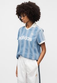 Φωτεινό γαλάζιο ριγέ τοπ adidas με λευκό γιακά, διακοσμημένο με μεγάλο λευκό λογότυπο. Συνδυασμένο με λευκό παντελόνι με μαύρες πλευρικές ρίγες.