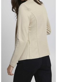 Beżowa marynarka o dopasowanym kroju, z gładką teksturą, skośnymi szwami i detalami z guzikami. Połączona z ciemnymi spodniami dla kontrastu.