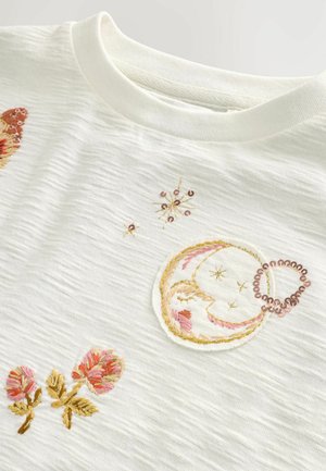 Valge tekstuuriga T-särk, millel on mitmevärvilised tikitud kujundid, sealhulgas lilled, taevalikud mustrid ja dekoratiivsed sequins.