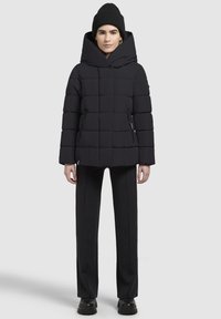 Schwarze Puffjacke mit großer Kapuze, gestepptem Design und Reißverschlusstaschen. Getragen mit schwarzen Hosen und einer schwarzen Mütze. Stiefel mit dicker Sohle.
