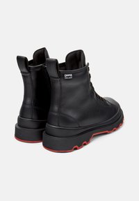 Un paio di stivali neri in pelle alla caviglia con suole rosse, zip laterale, lacci e etichetta "Camper Hydrashield" sulla caviglia esterna.