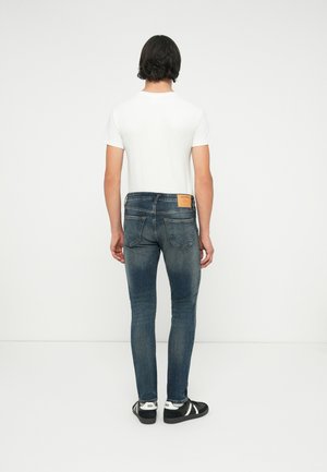 Muškarac tamne kose u bijeloj majici kratkih rukava, uskim plavim trapericama i crnim tenisicama stoji okrenut prema jednobijelom zidu.