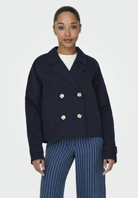 Giacca navy doppiopetto con bottoni dorati, colletto strutturato e due tasche laterali. Pantaloni a righe verticali, navy e bianchi.