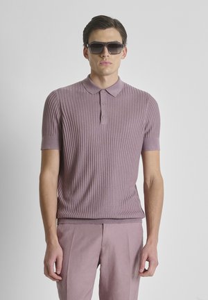 Homme portant un polo violet côtelé, un pantalon violet assorti et des lunettes de soleil foncées sur un fond gris uni.