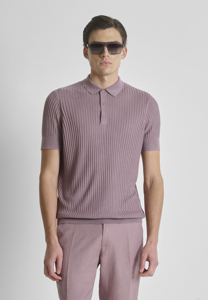 Hombre con camiseta tipo polo morada de canalé, pantalones morados a juego y gafas de sol oscuras sobre un fondo gris liso.