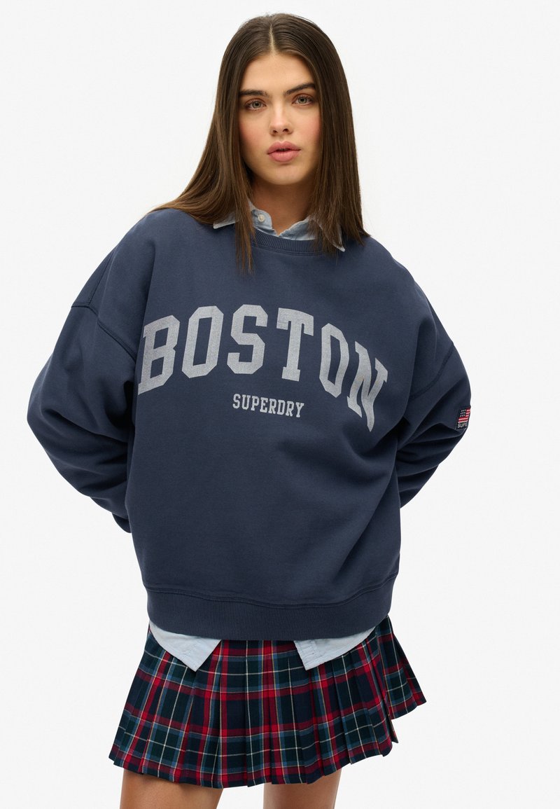 Sudadera extragrande azul marino con "BOSTON" y "SUPERDRY" en gris claro. Usada sobre una camisa de cuello azul claro, combinada con una falda a cuadros.