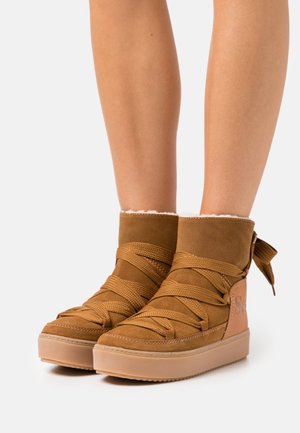 Hnědé zimní boty z semiše s tlustými křížovými tkaničkami a gumovou podrážkou, nošené na bosých nohách na bílém pozadí.