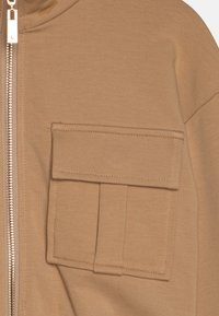 Sweatshirt beige zippé avec une texture douce, doté d'une grande poche à rabat à l'avant et d'une fermeture éclair en métal argenté sur un côté.
