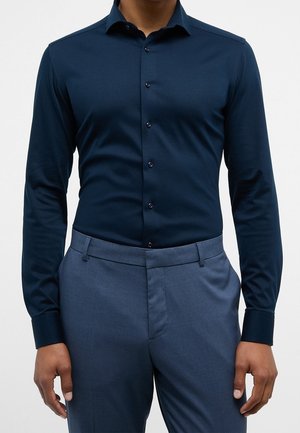 Mann trägt ein eng anliegendes dunkelblaues Hemd mit Knöpfen und hellblaue, maßgeschneiderte Hose, steht mit entspannten Armen an den Seiten.