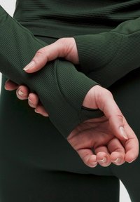 Ręce splecione w nadgarstkach, ubrane w ciemnozielone, prążkowane długie rękawy z otworami na kciuki, na tle ciemnego materiału.
