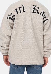 Beige fleecejacka med en texturerad yta, med svart broderad text på ryggen och ärmarna. Avslappnad passform med rundad nederkant.