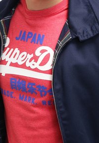 Maglietta rossa in cotone con testo blu "Superdry" e "JAPAN", indossata sotto una giacca con zip blu navy con fodera a quadri.