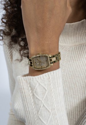 Náramkové hodinky ve zlaté barvě obdélníkového tvaru s diamantovými akcenty na zápěstí osoby, která nosí krémový svetr s copánkovým vzorem.