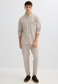 Camisa beige con textura y dos bolsillos en el pecho, combinada con pantalones cónicos a juego, usada con zapatos casuales marrones.