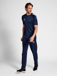 Čovjek stoji, nosi tamnoplavu sportsku odjeću s bijelim cik-cak detaljima i crne tenisice, gleda u stranu na jednobojnoj pozadini.