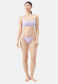 Frau, die barfuß steht und ein passendes Set aus lilafarbener BH und Stringunterwäsche trägt, schaut leicht zur Seite vor einem schlichten Hintergrund.