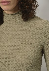 Olivengrünes, strukturiertes Strickoberteil mit hohem Kragen, das ein Rautenmuster aufweist und weiche, leicht erhabene Fasern für zusätzliche Dimension hat.