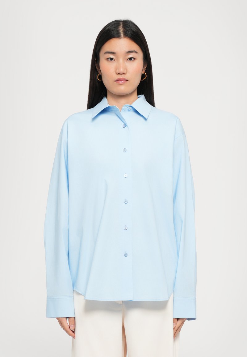 Helles blaues Button-Up-Hemd mit Kragen, langen Ärmeln und lockerer Passform. Verfügt über eine subtile Textur und Knöpfe entlang der Vorderseite.