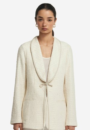 Weißer strukturierten Blazer mit Schalkragen, Schleifenverschluss und zwei vorderen Taschen. Der Stoff hat einen dezenten Glanz und ein gewebtes Muster.