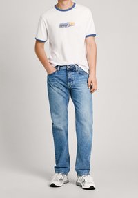 Bílé tričko s modrým lemováním u výstřihu a grafickým potiskem, doplněné světle modrými džíny s rovnými nohavicemi a bílými teniskami.