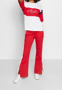 Camiseta deportiva Fila roja y blanca con el logo en el centro, combinada con pantalones rojos que tienen franjas laterales blancas y aberturas en el dobladillo. Zapatillas blancas.