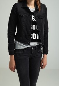 Chaqueta de mezclilla negra con un diseño ajustado, botones frontales y bolsillos en el pecho. Llevada sobre una camiseta gris con un texto gráfico en negrita, de color negro.