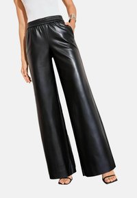 Platām kājām melnas ādas bikses ar elastīgu jostasvietu un sānu kabatām, demonstrējot gludu, spīdīgu tekstūru un brīvu siluetu.