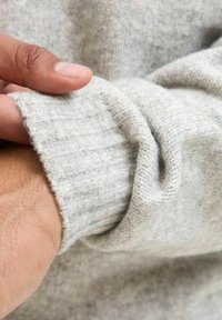 Lys grå strikket sweatermanchet med ribbet tekstur, holdt af en hånd, der fremviser blødhed og let stræk.