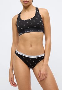 Μαύρο αθλητικό σουτιέν από βαμβάκι και αντιστοιχισμένο μπικίνι, και τα δύο με επαναλαμβανόμενο λογότυπο «CK». Το σουτιέν έχει φαρδιές τιράντες· ελαστικές ζώνες μέσης σε αντίθετα χρώματα.