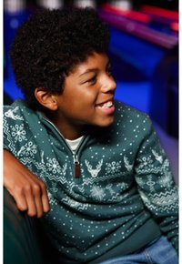 Grön zip-up tröja med vita snöflinge- och renmönster, bärs över en ljus skjorta. Kort, lockigt mörkt hår. Avslappnad miljö.