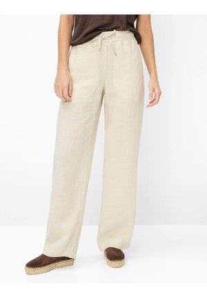 Person trägt beige weite Hosen mit Kordelzug und braune Espadrille-Schuhe und steht vor einem schlichten weißen Hintergrund.