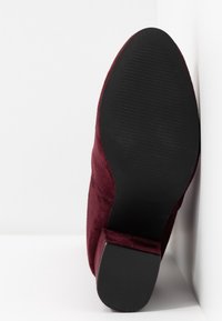 Semelle et talon d'une bottine cheville en daim bordeaux reposant contre une surface blanche, montrant une semelle extérieure en caoutchouc noir texturée pour l'adhérence.