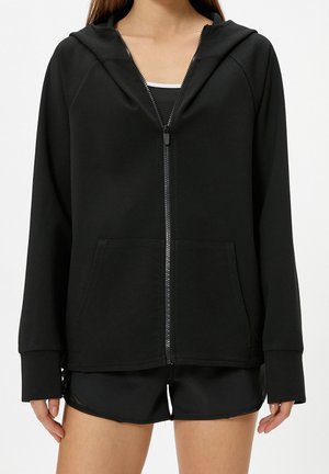 Vrouw met een zwarte zip-up hoodie met voorzakken en zwarte sportshorts, getoond van nek tot midden dij tegen een witte achtergrond.