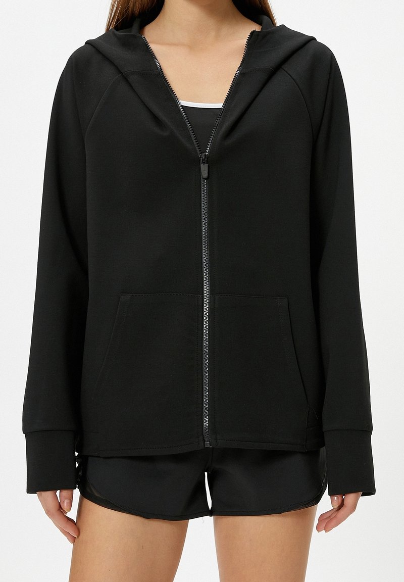 Femme portant un sweat à capuche noir zippé avec poches avant et un short de sport noir, visible du cou jusqu'à mi-cuisse sur un fond blanc.