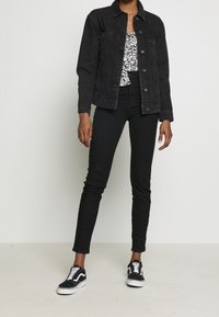 Persona con chaqueta de mezclilla negra, jeans skinny negros, zapatillas en blanco y negro, y una blusa floral en blanco y negro, de pie contra una pared blanca.