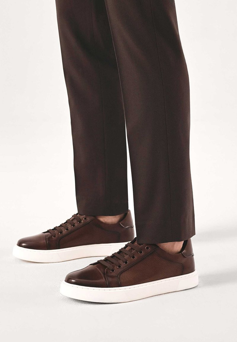 Barna bőr sneaker texturált panellel, fehér gumitalppal és kerek orral. Fekete fűző és fém szemek, sötét nadrággal szemben.