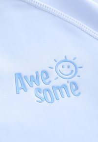 Witte stof met een geborduurd blauw ontwerp met de tekst "Awe some" en een glimlachende zon boven de tekst. Gladde textuur.