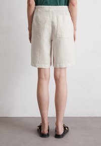 Pantalones cortos de lino en beige claro con un ajuste relajado, de longitud por encima de la rodilla y un solo bolsillo trasero. Combinados con sandalias negras.