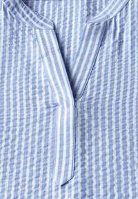 Blouse en coton rayée bleue et blanche avec un col en V, présentant des rayures alternées verticales et horizontales et une finition texturée.