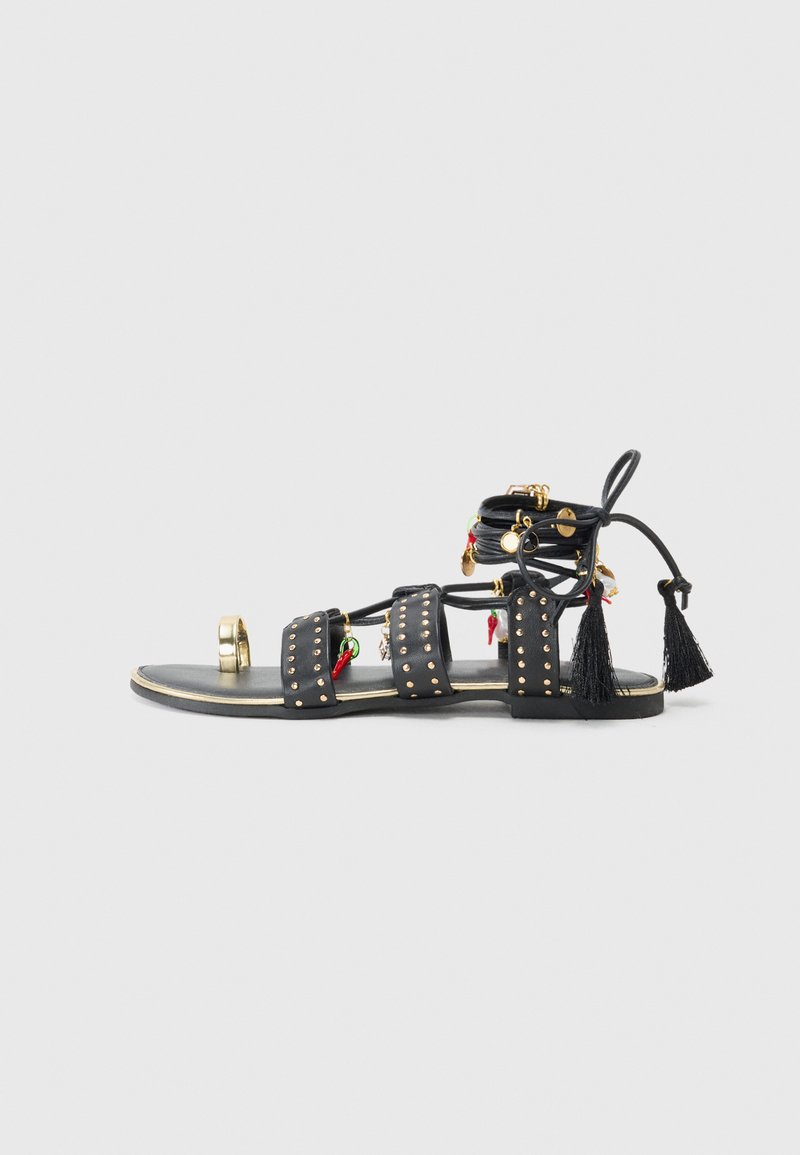 Sandali in pelle nera con tre cinturini larghi impreziositi da borchie dorate, nappine decorative e charm rossi e verdi. Suola piatta con un accento dorato.