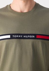 Muž má na sobě olivově zelené tričko s crewneck výstřihem, na kterém je černý, bílý a červený pruh přes hruď s logem "Tommy Hilfiger".