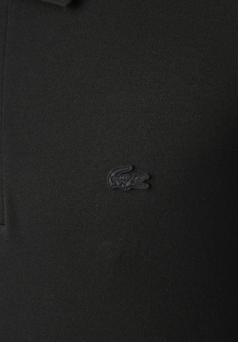 Chemise polo noire en tissu texturé, ornée d'un petit logo crocodile brodé de couleur sombre sur le côté gauche.
