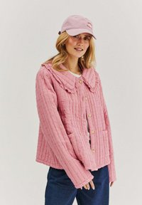 Světle růžová prošívaná bunda s velkým límcem, předními knoflíky a dvěma kapsami. Textura má jemný tečkovaný vzor.