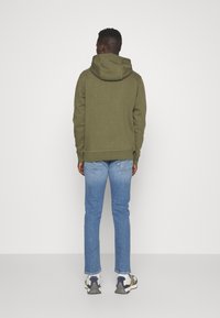 Sudadera verde oliva con capucha, puños y dobladillo acanalados, combinada con vaqueros de azul claro y zapatos deportivos grises, vista desde la parte posterior.