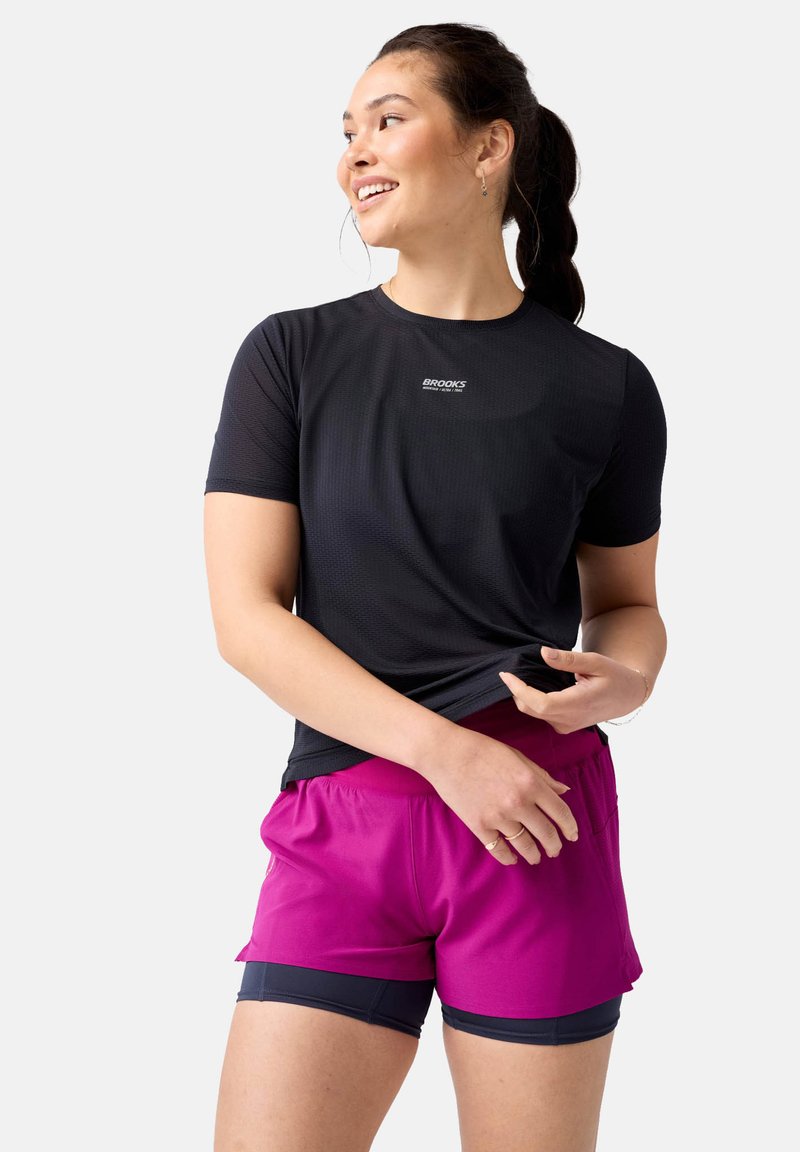 Camiseta deportiva negra de manga corta con tejido texturizado y el logo "Brooks", combinada con pantalones cortos rosa vibrante que cuentan con una cinturilla elástica.