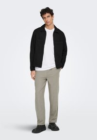 Μαύρο σακάκι με δύο μπροστινές τσέπες, λευκό μπλουζάκι, ανοιχτό γκρι παντελόνι και μαύρες μπότες, στέκεται μπροστά σε απλό λευκό φόντο.