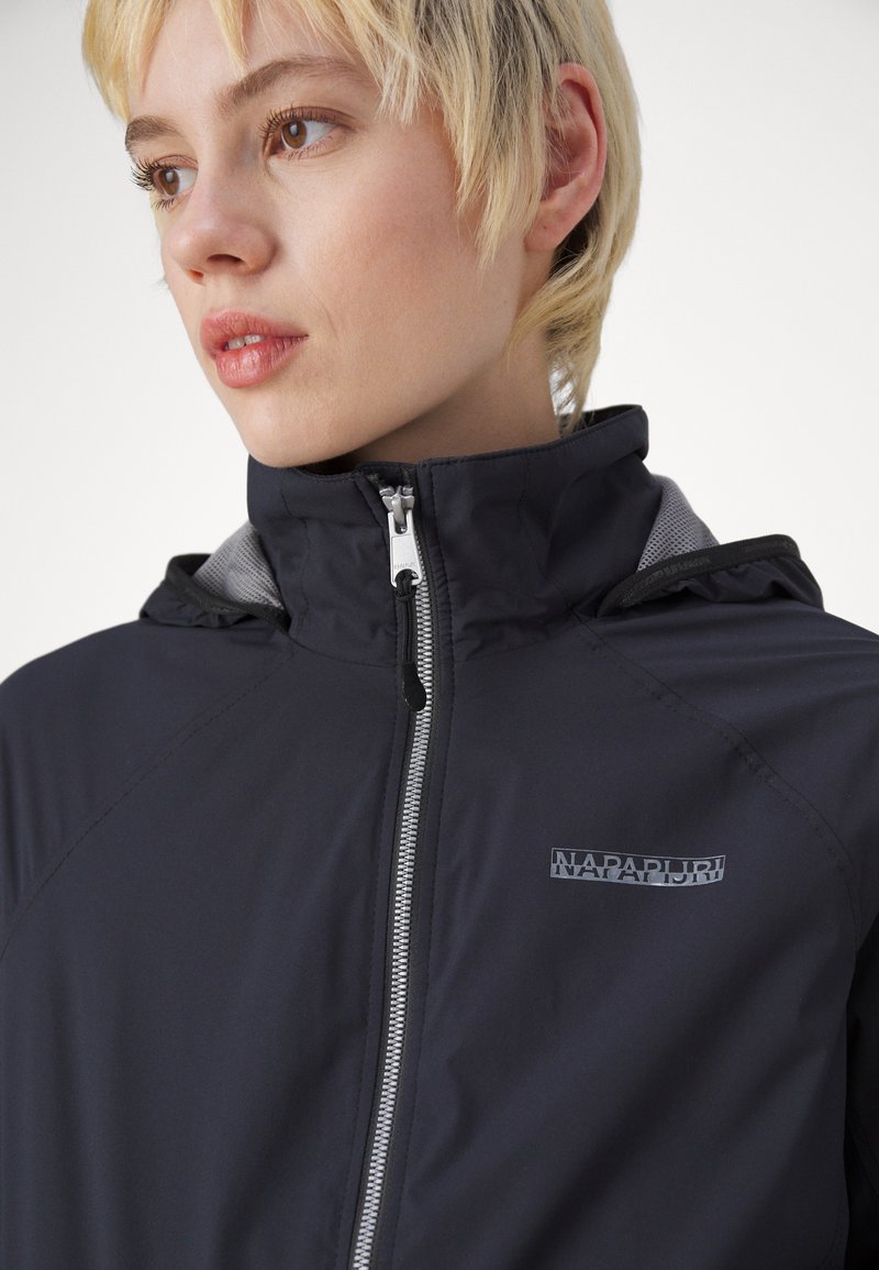 Giovane con capelli biondi corti che indossa una giacca nera Napapijri con zip, colletto alto e cappuccio.